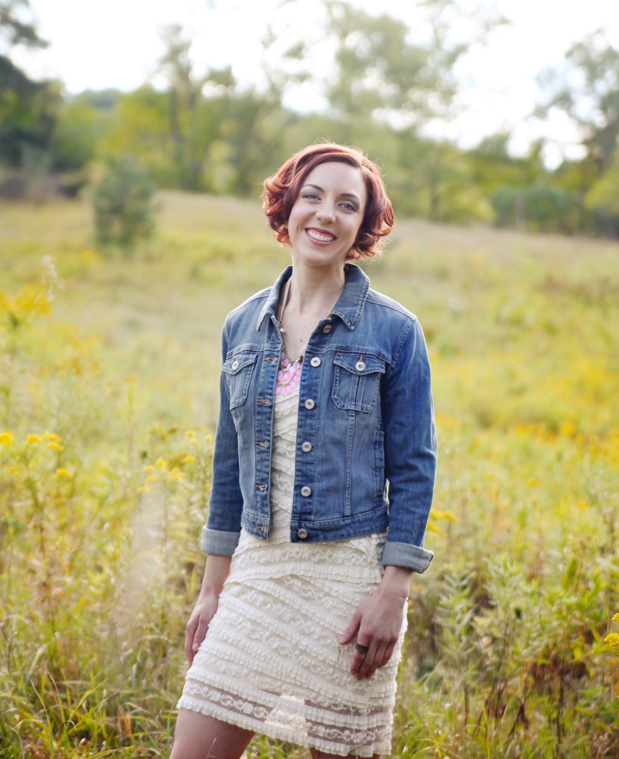 Katy standing in a field of flowers, wearing a lace dress with a jean jacket on top