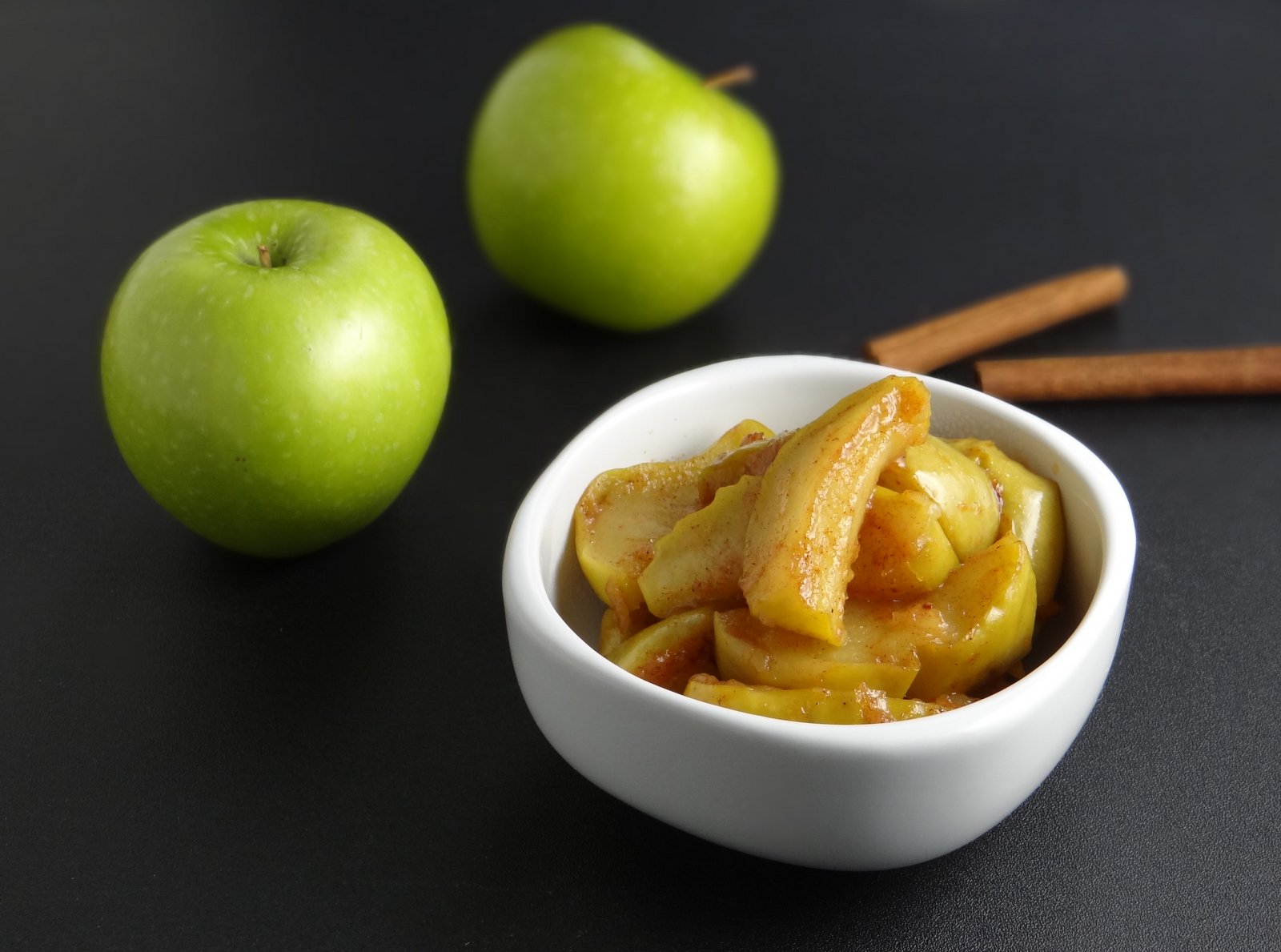 fried apples served in a white bowl with 2 raw apples and 2 cinnamon sticks in the background