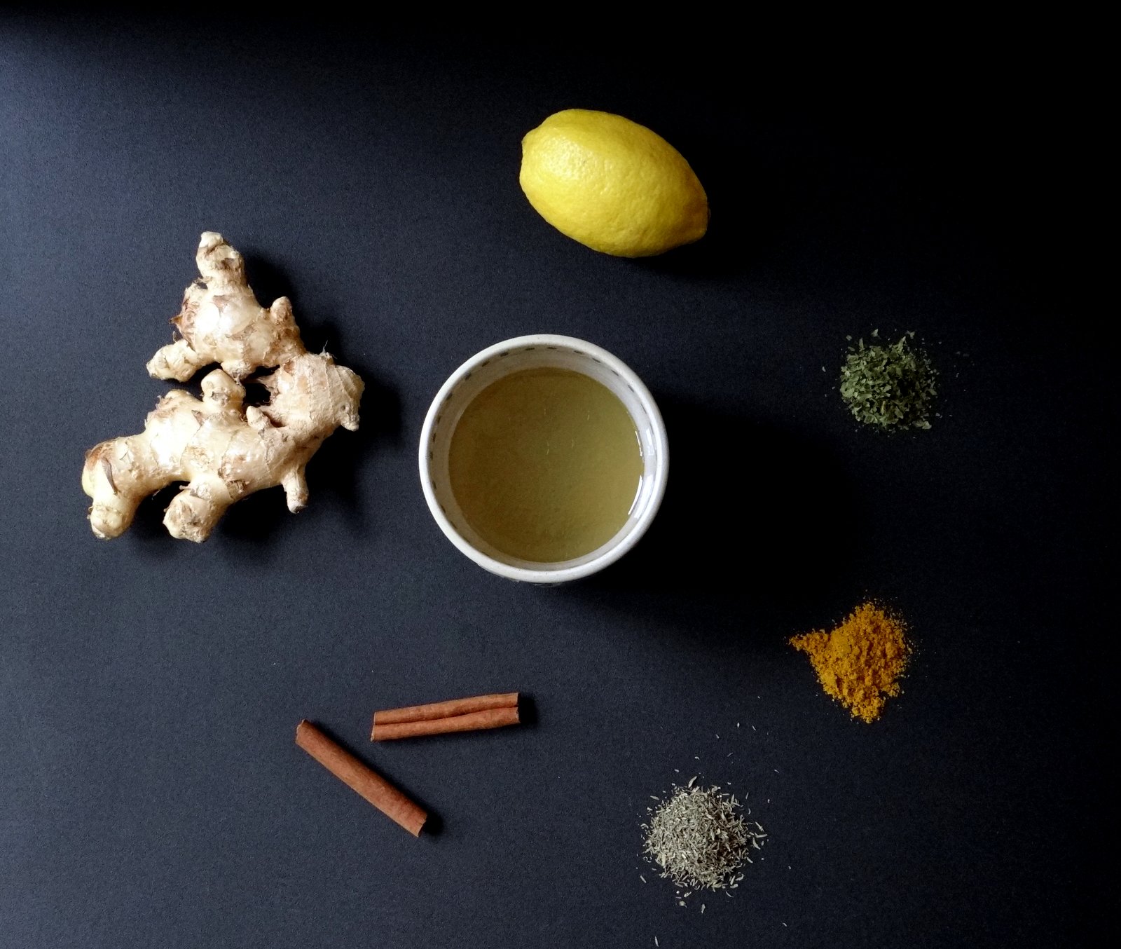 ingredients on black background: cup of broth, ginger, whole lemon, cinnamon sticks, and herbs