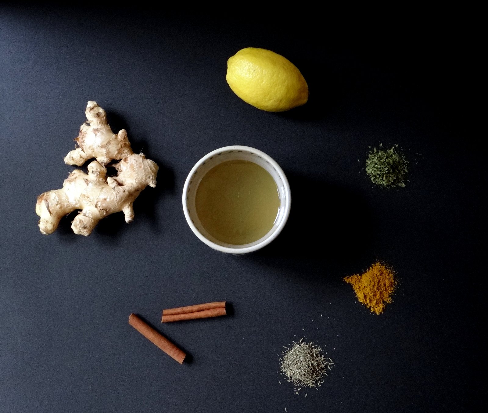 ingredients on black background: cup of broth, ginger, whole lemon, cinnamon sticks, and herbs