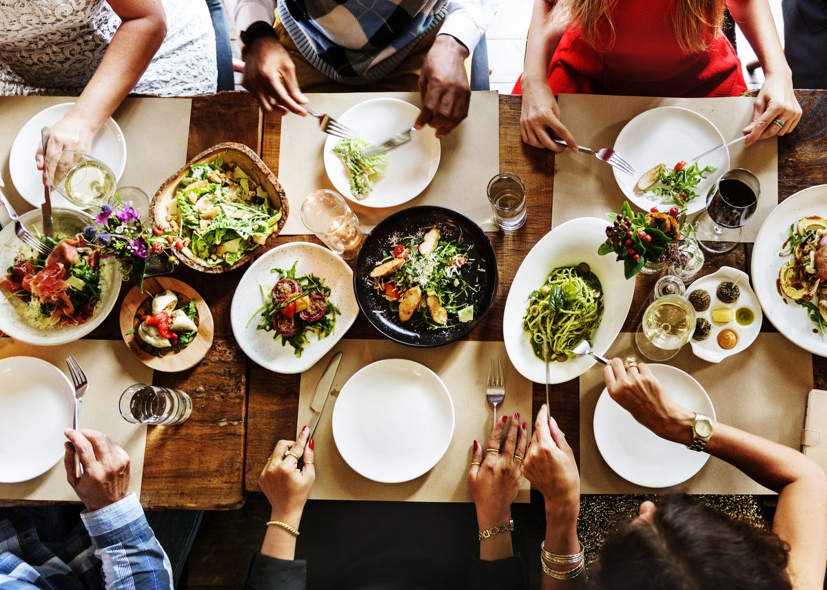 Top view of dinner table with food in the center, and people reaching to fill their plates.