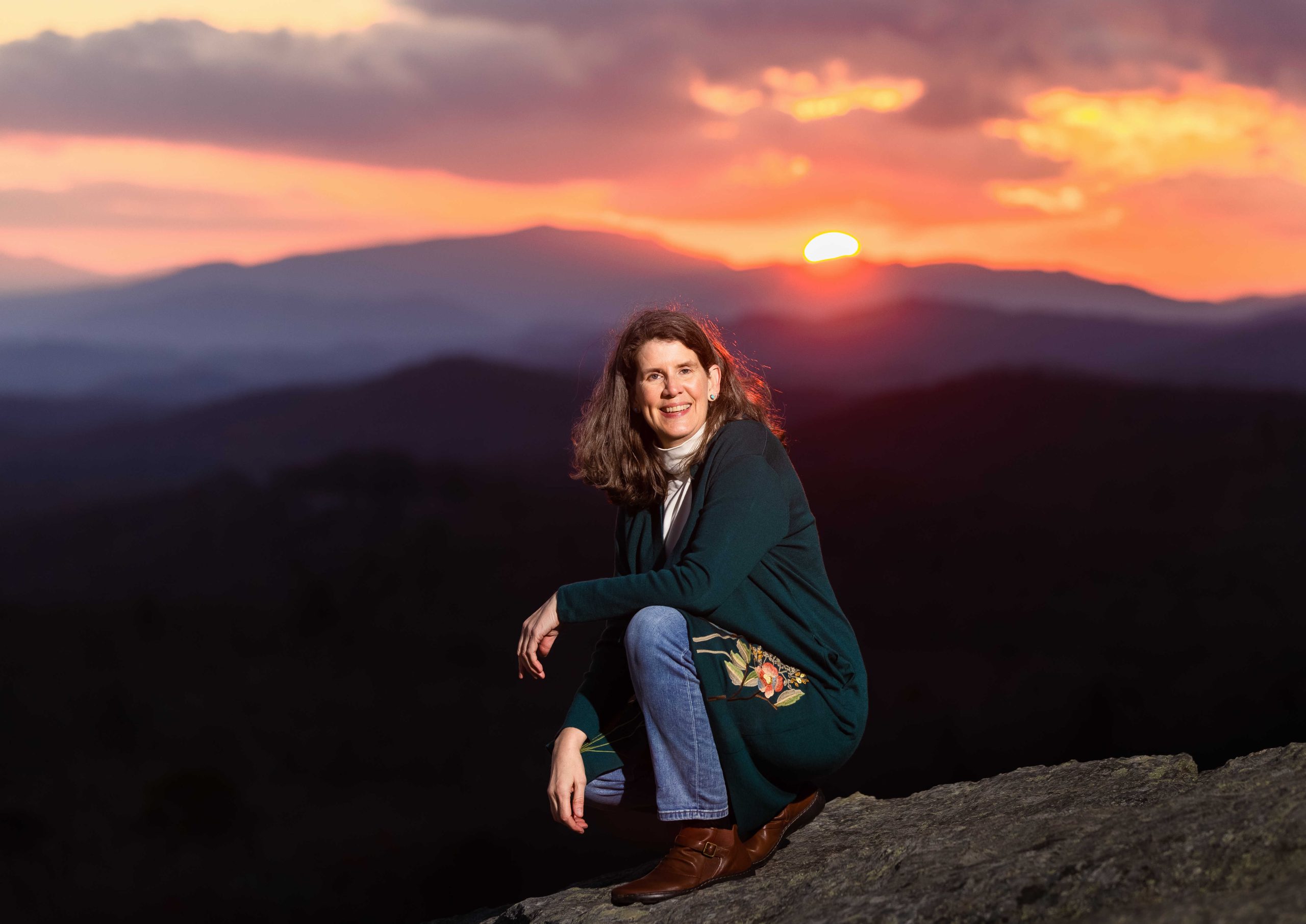 author squatting with a mountain vista behind her at sunset
