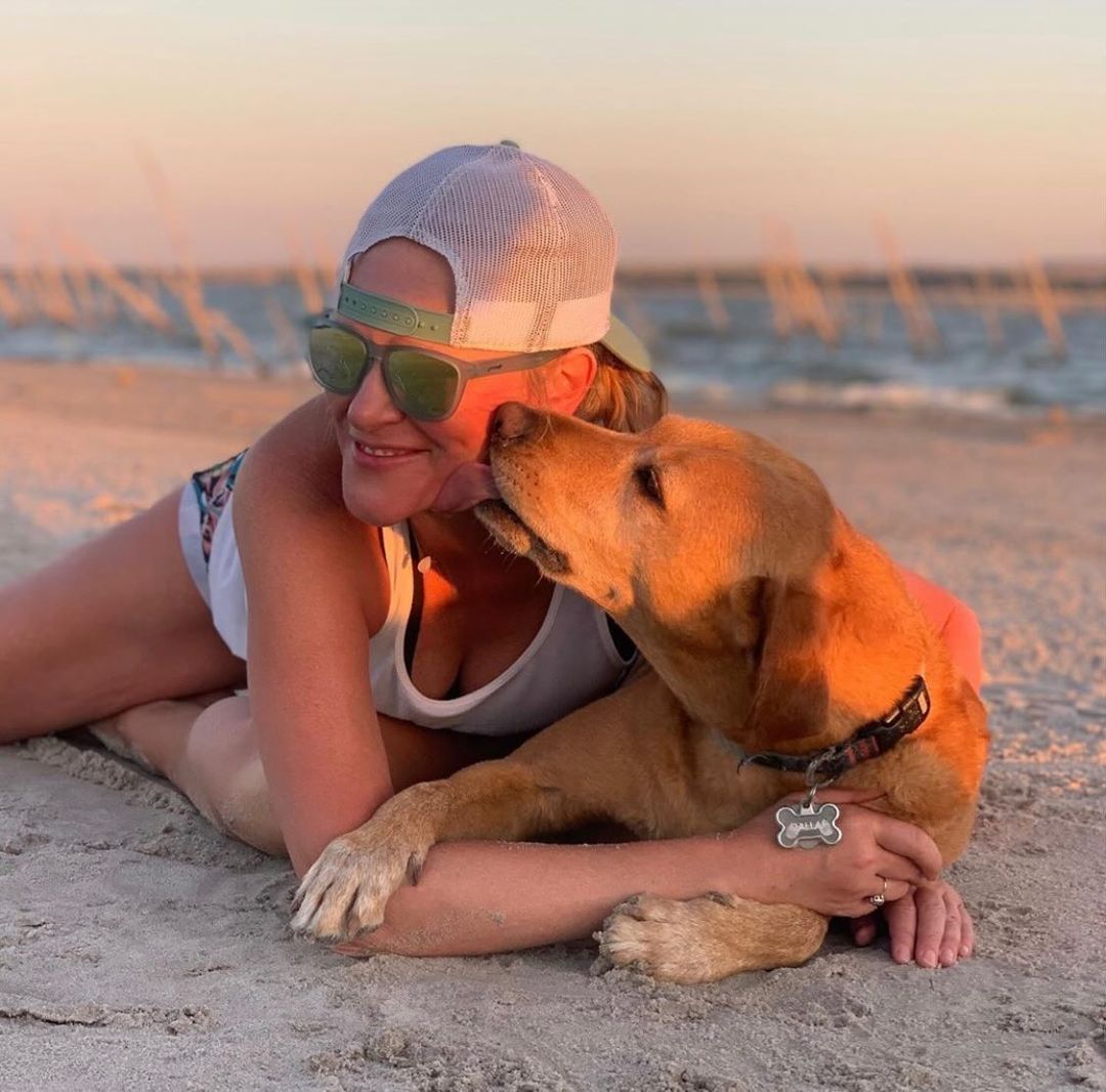 Lora Marie and her dog lying on the beach as her dog licks her face