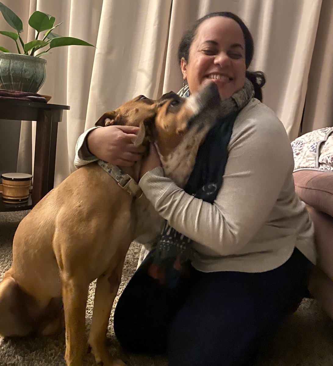 Melissa laughing as she kneels on the floor, hugging her dogs as they try to lick her face